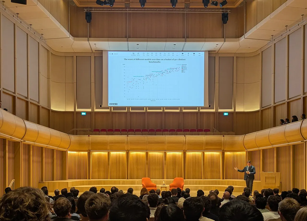 Anthropic 联合创始人 Jack Clark 在牛津大学的礼堂里做关于 AI 的演讲，大屏幕上是各模型评测分数随时间上升的图表